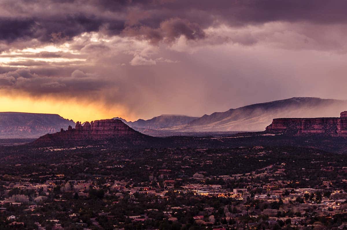 View of Sedona during monsoon season with dark clouds, rain bands, and glowing sky over red rock formations