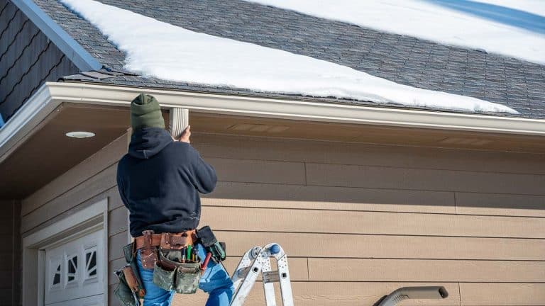 A roofer working on a gutter beneath a snow-covered roof in Prescott. From Hahn Roofing's "5 Winter Roofing Tips to Keep Your Prescott Home Safe."